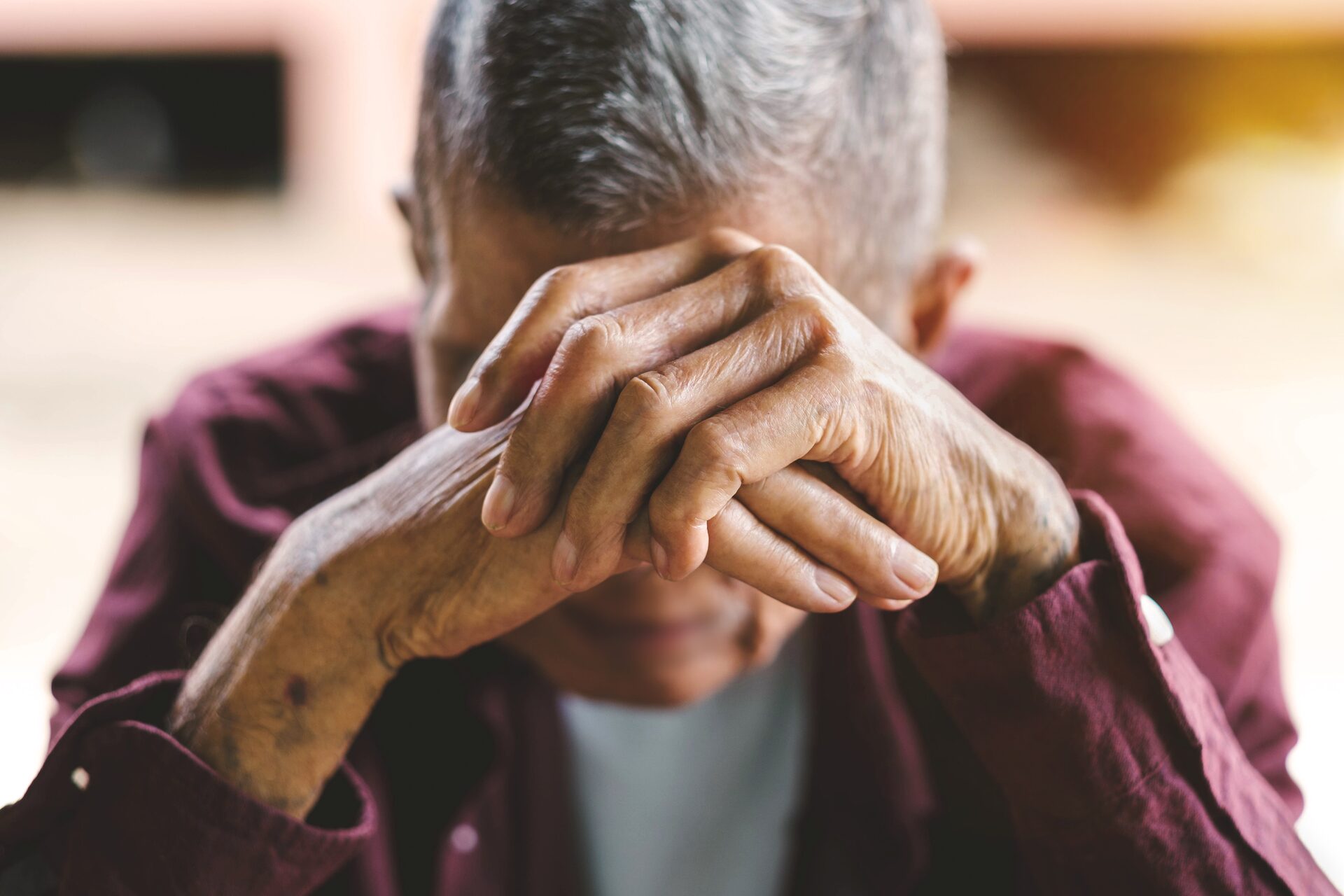 An elderly person seated, head lowered into folded hands
